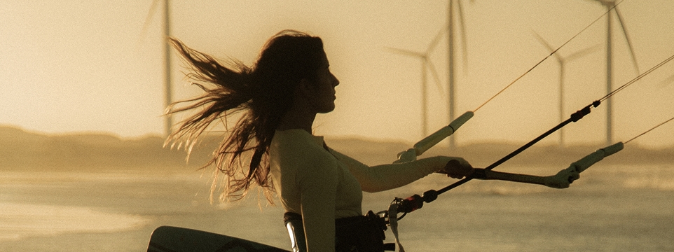 Woman kitesurfing at sunset, with wind turbines in the background.