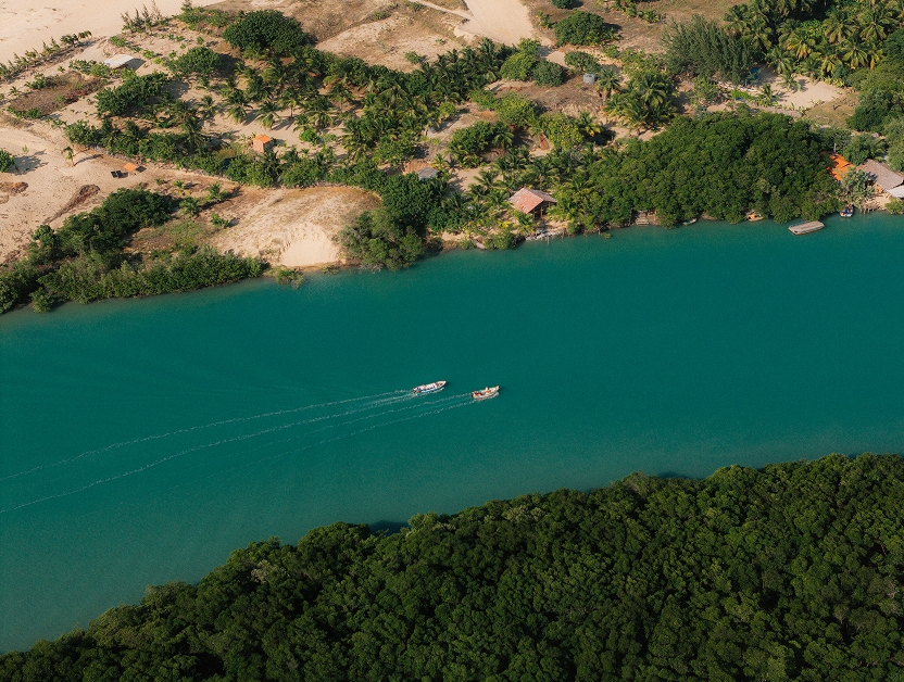 Aerial view of a blue-green river with two boats, between vegetation and dunes.