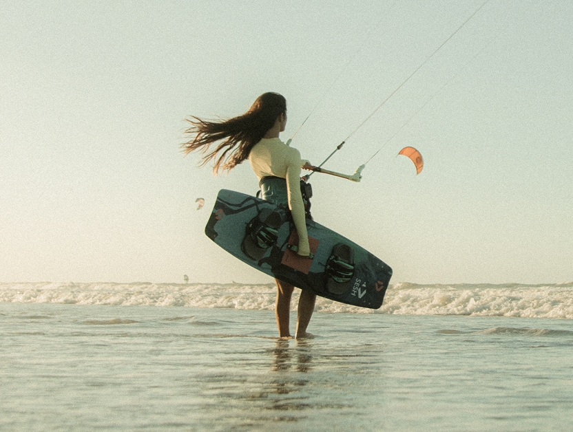Woman seen from behind holding a kitesurf board at the water's edge