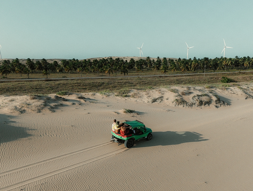 Green buggy with people crossing the dunes, with palm trees and wind turbines in the background