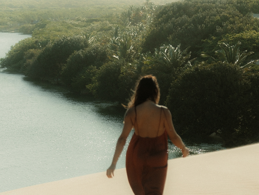 Woman in a red dress seen from behind on a dune, with a river and vegetation in the background